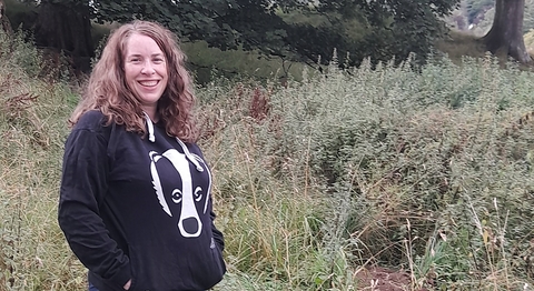 A smiling woman in a hoodie with a badger on it, standing in front of a badger sett