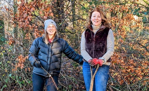 Two women standing outdoors, wrapped up warm. One is holding a shovel. 