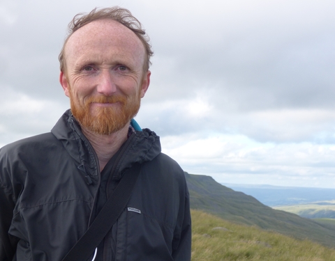 A man in a waterproof jacket standing in front of a hilly landscape