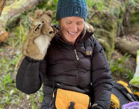 A woman sitting in a forest with a wolf hand puppet on her hand