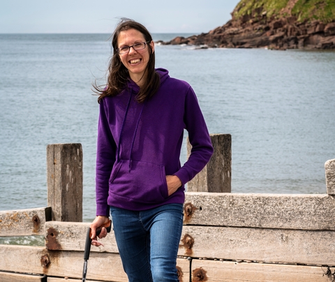 A woman stands on the shore at the beach, in front of the sea and a section of cliff. 