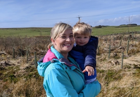 A woman in front of fields on a sunny day, holding a young child.