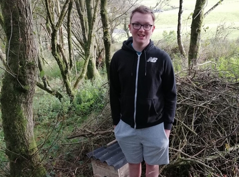 A young man standing in the woods next to a wooden hedgehog house