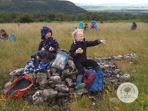 Two children eating their packed lunches on a rock in the middle of a field, with other children in the background