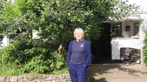 A woman standing in front of a building and foliage, with her hands in her pockets