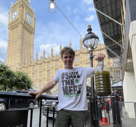 A man in a #SaveTheMere t-shirt, holding up a big bottle of grim-looking green water