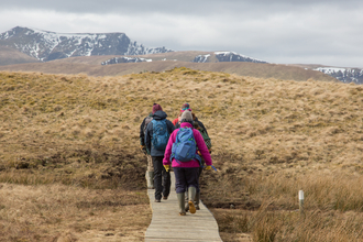 Group of walkers crossing wooden boardwalk over Eycott Hill Nature Reserve wetland with snow topped fells in background