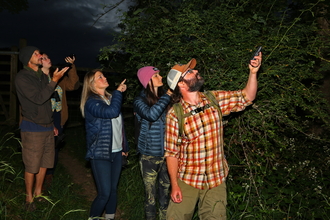 A group of people standing in a dark forest, holding up bat detectors.