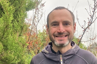 A man smiling into the camera in front of some trees