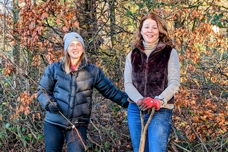 Two women standing outdoors, wrapped up warm. One is holding a shovel. 