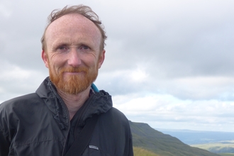 A man in a waterproof jacket standing in front of a hilly landscape