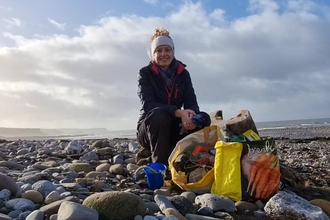 A woman crouching on a stony beach with bags of rubbish. 
