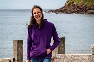 A woman stands on the shore at the beach, in front of the sea and a section of cliff. 