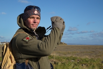 A man standing on a beach holding binoculars and looking into the camera