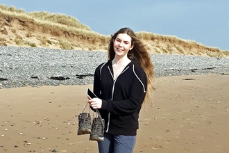 A young woman on a sandy beach, holding a pair of walking boots. 