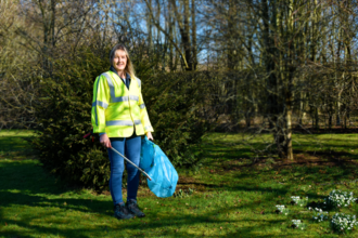 A woman in a hi-vis jacket holding a plastic bag and a litter picking tool in front of trees