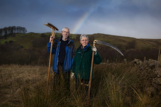 A man and a woman standing in a field, holding farming tools, with a rainbow behind them