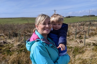 A woman in front of fields on a sunny day, holding a young child. 