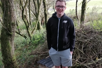 A young man standing in the woods next to a wooden hedgehog house