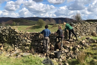 Three people leaning on a dry stone wall, looking across at the hills beyond, underneath a blue sky.