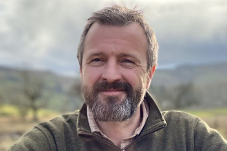 A man with a beard, wearing a zip-up fleece, leans on a gate in front of a field. 