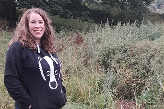 A smiling woman in a hoodie with a badger on it, standing in front of a badger sett