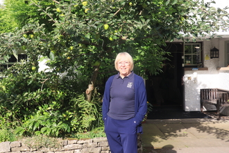 A woman standing in front of a building and foliage, with her hands in her pockets