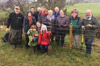 A group of people, from children to older people, leaning against a wire fence with tree planting equipment