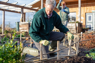 A man in a green fleece and gardening gloves kneeling near a small fence in a wildlife garden