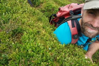 A man in a cap and carrying a rucksack crouches next to a bush, smiling. 