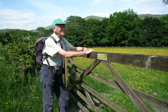 A man wearing a cap and rucksack leans against a wooden five-barred gate, in front of a field full of wildflowers. 