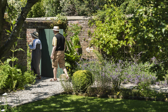 Photo shows two people walking through a green door in a stone wall. In the foreground is a grass lawn with a flowerbed full of plants, shrubs and flowers
