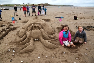 Two children in front of a large sand sculpture of a crab on the beach.