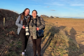 Two young women standing on beach with long-lens cameras and shadows behind them