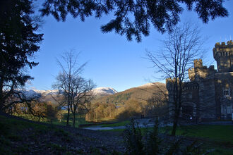 A landscape photo with bushes and trees in the foreground, part of Wray Castle on the right side, and fells capped with snow in the background