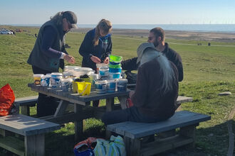 A group of four people seated on a picnic bench organising buckets, with the sea in the distance.