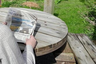 Someone sitting at a picnic table reading a newspaper.