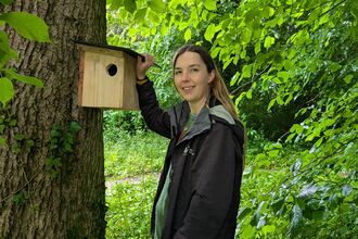 Ella Blundell holding up a red squirrel nesting box against a tree in a woodland setting