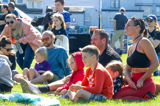 A group of adults and children sitting on the grass looking off-camera at a performance, with stalls in the background