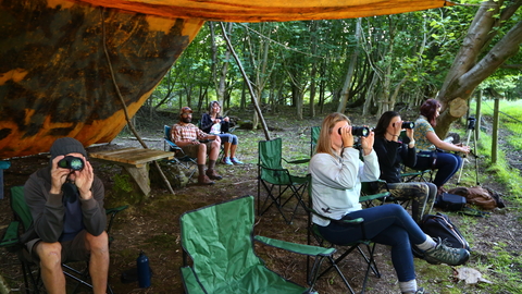 People sitting under a canvas hide, looking at wildlife through binoculars