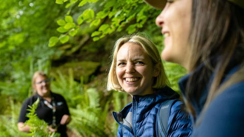Three people in outdoor gear standing in a leafy woodland, smiling