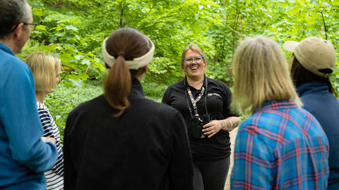 A group of people standing around a smiling group leader, against a woodland backdrop