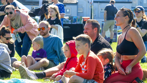 A group of adults and children sitting on the grass looking off-camera at a performance, with stalls in the background