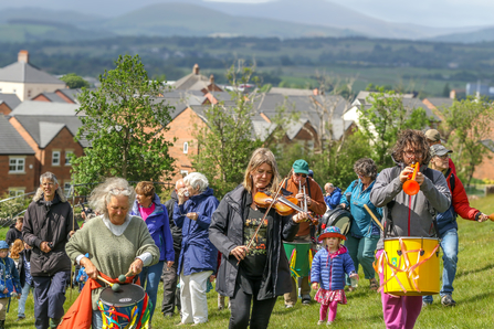 Group of people walking up hill playing musical instruments with fells in background