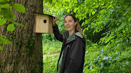 Ella Blundell holding up a red squirrel nesting box against a tree in a woodland setting
