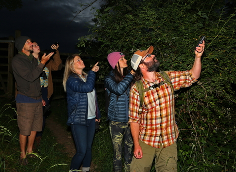 A group of people standing in a dark forest, holding up bat detectors.